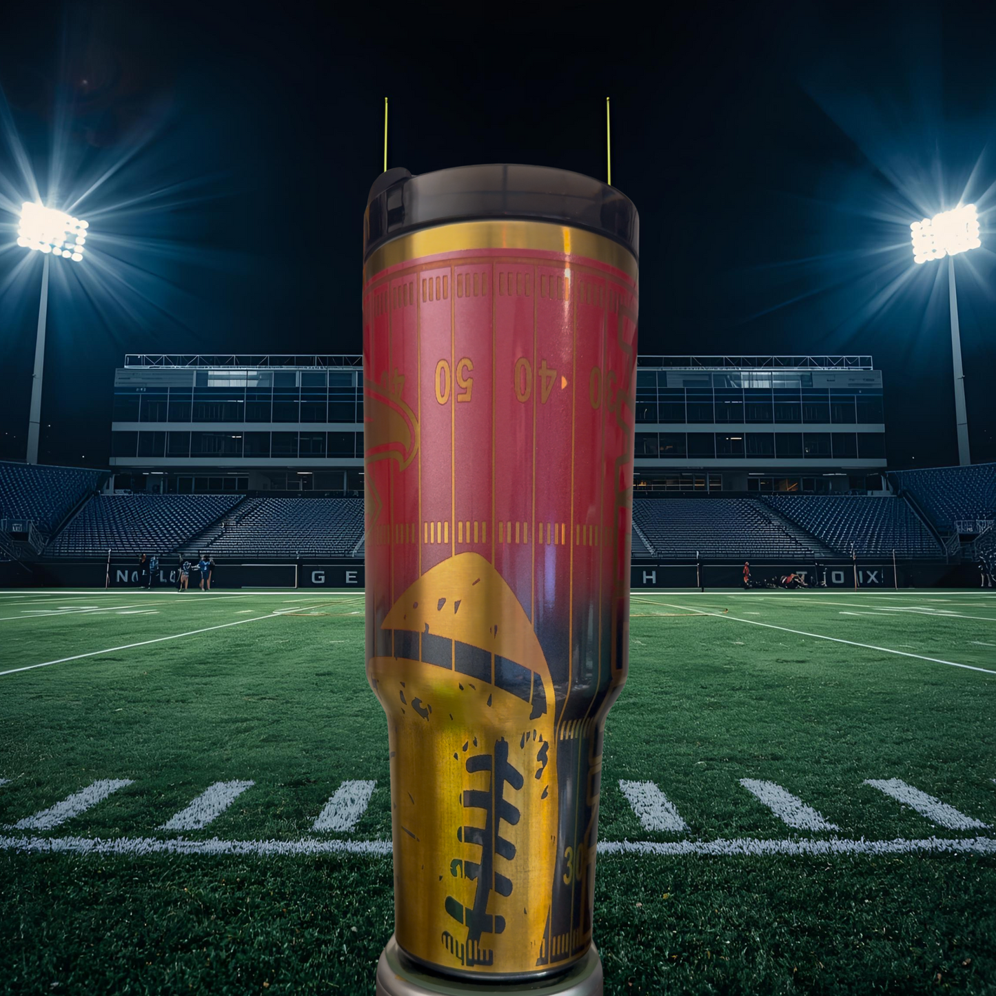 A large, colorful tumbler with a football design on it, placed on a football field with stadium lights in the background.